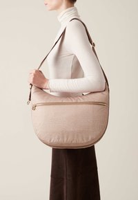 Pink textured shoulder bag with a curved shape, featuring a gold zipper and a brown strap, modeled on a neutral backdrop.