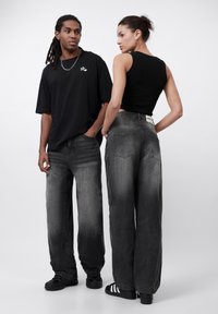 Svart oversized t-skjorte, grå vide jeans. Glatt stoff med en falmet finish. To modeller som viser bakfra, minimal merkevare på jeansene.