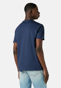 Joven de pie de lado, vistiendo una camiseta de manga corta azul marino sin estampados y jeans de mezclilla azul claro, contra un fondo liso.