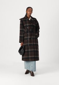 Abrigo trinchera de doble botonadura en cuadro oscuro, con cintura ceñida por un cinturón, cuello con muescas y grandes botones. Combinado con una falda de mezclilla y botas marrones.