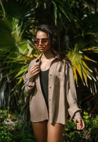 Camicia button-up marrone chiaro con maniche arrotolate, indossata su un costume da bagno nero a fantasia, ambientata tra la vegetazione tropicale verde.
