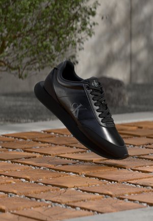 Basket en cuir noir avec logo, flottant au-dessus d'un sol en carreaux de bois en extérieur près d'un arbre et d'un mur en béton.