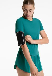 Žena v tyrkysovom športovom oblečení si nastavuje čierny smartphone armband na hornej časti ľavého ramena na bielom pozadí.