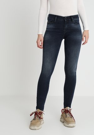 Kvinde iført mørkeblå skinny jeans, beige chunky sneakers med røde snørebånd og en tætsiddende langærmet hvid top, stående på en ensfarvet baggrund.