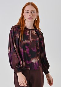 Blusa fluida a maniche lunghe di colore viola scuro con motivi astratti. Il collo a forma di occhiello e i polsini arricciati aggiungono dettagli. Tessuto leggero.