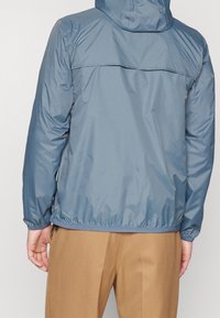 Leichte blaue Jacke mit Kapuze, elastischen Bündchen und Saum. Verfügt über ein Belüftungsnetz am Rücken und eine glatte, glänzende Textur.