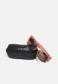 Rosa solbriller med gjennomsiktig ramme og brune linser, hvilende på et svart hardt etui med glidelås og hvit "vogue"-logo.