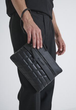 Bolso de cuero negro con patrón acolchado, forma plana, cierre superior con cremallera y correa de muñeca desmontable. Superficie texturizada con acentos lisos.