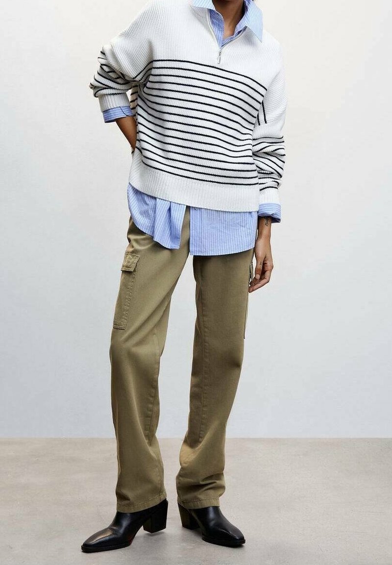 Maglione bianco a righe con zip, indossato sopra una camicia button-up azzurro chiaro, abbinato a pantaloni cargo khaki e stivali neri con tacco.