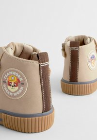 Stivaletti beige con accenti marroni, suole in gomma testurizzata e dettagli cuciti. Presentano una toppa rotonda con un cane dei cartoni animati e uno slogan.