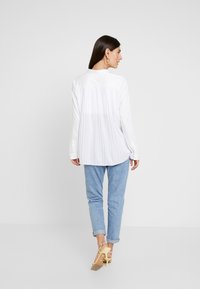 Weiße Bluse mit langen Ärmeln und plissiertem Rücken, aus weichem Stoff, mit abgerundetem Saum, kombiniert mit hellblauen hochgekrempelten Jeans und gelben Absätzen.
