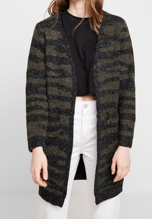 Femme portant un cardigan ouvert duveteux à motif camouflage, un top court noir et un pantalon blanc taille haute avec fermeture à boutons.
