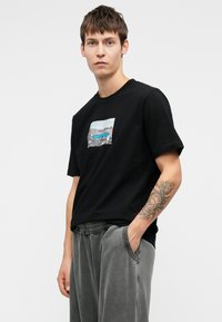 Tânăr îmbrăcat cu un tricou grafic negru și pantaloni gri, cu o mână în buzunar, tatuaj vizibil pe antebrațul drept, fundal neutru.