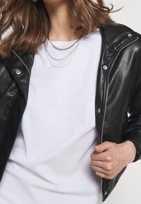 Černá kožená bunda s kovovými detaily, zipovým zapínáním a knoflíky, přehodnocená přes jednobarevné bílé tričko. Modelka drží bundu zavřenou.