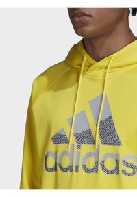 Felpa gialla con cappuccio regolabile, caratterizzata da un grande logo Adidas grigio con un motivo a pois sul davanti. Tessuto liscio.