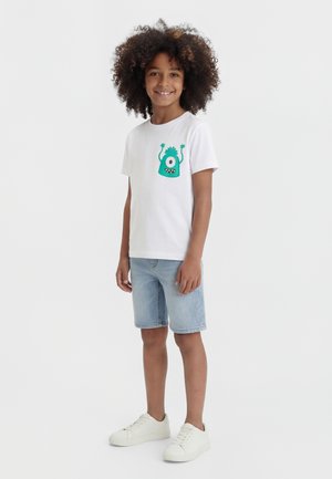 Kind met krullend haar draagt een wit T-shirt met een groene eenoog monster afbeelding, lichte denim shorts en witte sneakers, staand en glimlachend.
