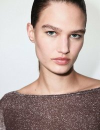 Portret osoby w błyszczącej brązowej bluzce na ramiączkach. Tło jest jasne, co podkreśla jednolity koloryt skóry oraz niebieskie oczy.