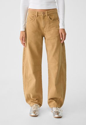 Persoon draagt wijd uitlopende beige broek, wit shirt met lange mouwen en zilveren veterschoenen, staand tegen een effen achtergrond.