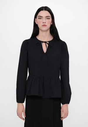 Black peplum blouse with long sleeves, keyhole neckline fastened by a tie, smooth texture, and gathered hem, worn over a pleated skirt.
