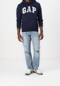 Homme portant un sweat à capuche GAP bleu marine, un jean déchiré bleu clair et des baskets blanches avec des rayures noires, se tenant contre un fond blanc.