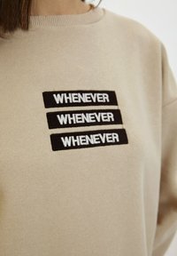 Felpa beige con tre toppe rettangolari nere ricamate con la scritta "WHENEVER" in bianco disposta in modo impilato.