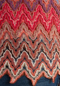 Häkeloberteil mit einem Zickzackmuster in Rot-, Korallen- und Beigetönen, mit einer strukturierten, offenen Webart und gewellten Kanten.