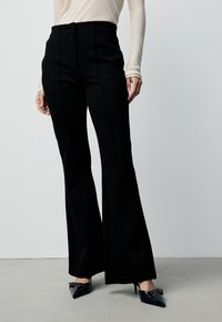Black flared pants with a high waist and straight shape. Smooth fabric with front seam detailing, paired with pointed black heels.
