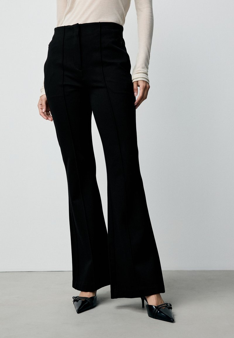 Black flared pants with a high waist and straight shape. Smooth fabric with front seam detailing, paired with pointed black heels.