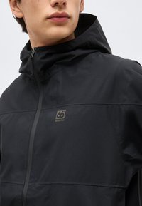 Jeune personne portant une veste noire à capuche fermée, avec un petit logo 66° North sur le côté gauche de la poitrine.