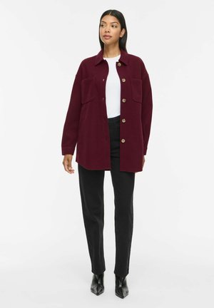 Femme portant une veste bordeaux boutonnée sur une chemise blanche, un pantalon noir et des bottes pointues noires, debout devant un fond uni.