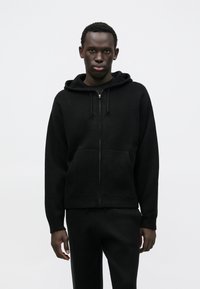 Czarna bluza z kapturem rozpinana na zamek, wykonana z miękkiego materiału, z kieszenią typu kangur, kapturem ze sznurkiem oraz ściągaczami przy mankietach. Do kompletu dopasowane czarne spodnie.