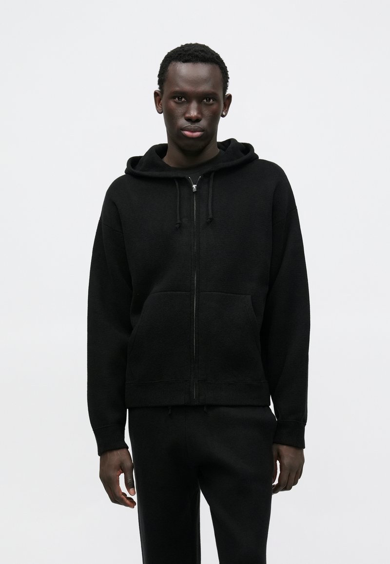 Czarna bluza z kapturem rozpinana na zamek, wykonana z miękkiego materiału, z kieszenią typu kangur, kapturem ze sznurkiem oraz ściągaczami przy mankietach. Do kompletu dopasowane czarne spodnie.