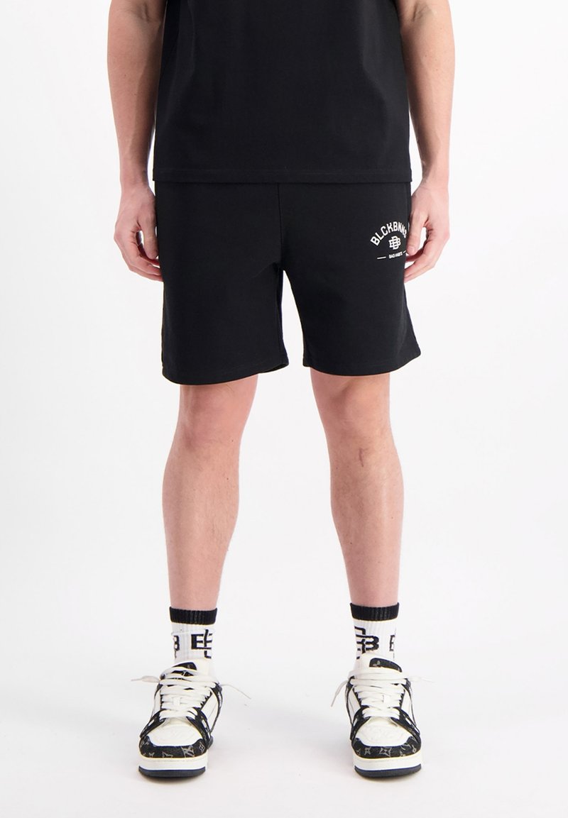 Zwarte shorts van een gladde stof, met een klein wit logo op het linkerpijp. Draag met zwarte sneakers en gemusterde sokken.