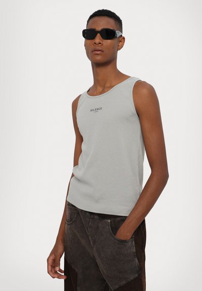 Grijze geribbelde tanktop met "SILENCE" in het zwart gedrukt, gedragen met donkerbruine, twee-kleurige broek en zwarte rechthoekige zonnebril.
