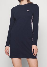 Mörkblå klänning med lång ärm, tillverkad av bomull, med kontrasterande ränder längs sidorna och en liten, broderad logotyp på bröstet.