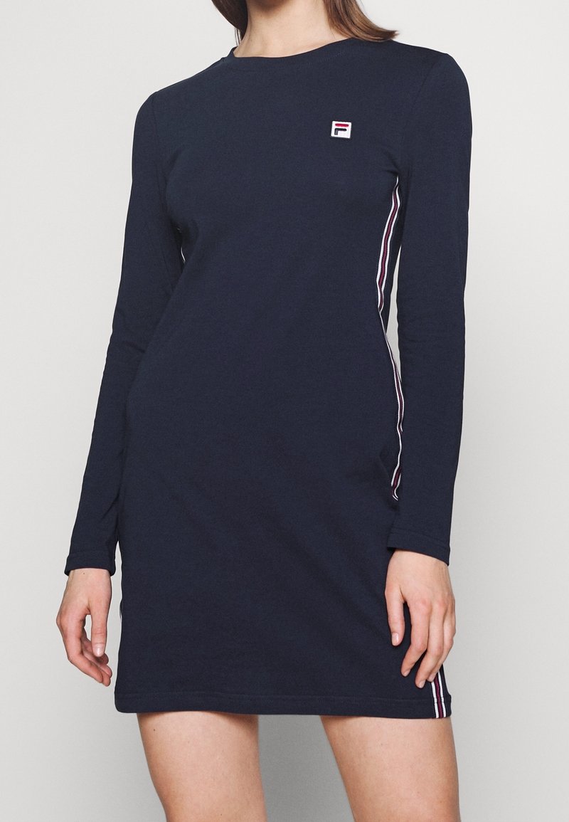Mörkblå klänning med lång ärm, tillverkad av bomull, med kontrasterande ränder längs sidorna och en liten, broderad logotyp på bröstet.