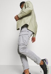 Jasnozielony długi rękaw z zaokrąglonym dołem, połączony z jasnoszarymi spodniami typu cargo z kieszeniami i detalem na suwak. Czarne trampki z fioletowymi akcentami.