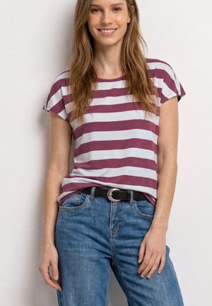 Ung kvinde med langt brunt hår iført en lilla og hvidstribet skjorte, blå jeans og et sort bælte, stående mod en hvid væg.