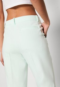 Femme portant un pantalon vert menthe clair ajusté et un top court blanc, une main dans la poche du pantalon, vue de dos.