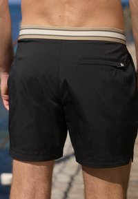 Svarta badshorts med randig, elastisk midjeresår och en bakficka med dragkedja. Slätt tyg med minimal textur.