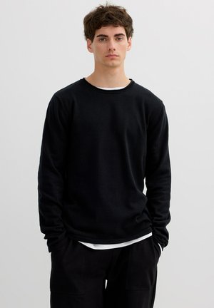 Joven con cabello corto y castaño, vestido con una camisa negra de manga larga sobre una camiseta blanca, de pie con las manos en los bolsillos contra un fondo liso.