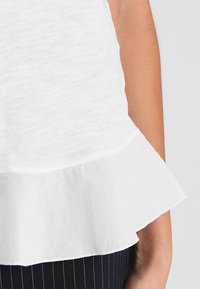 Top blanco con un dobladillo de volantes en capas, con una tela texturizada. El top tiene mangas cortas y un corte holgado, combinado con pantalones a rayas.