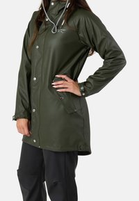 Femme portant un imperméable vert foncé avec des boutons-pression et un pantalon noir, debout avec une main sur la hanche.