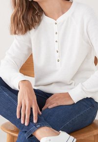 Femme assise sur une chaise en bois portant un haut boutonné à manches longues blanc, un jean bleu et des baskets blanches, avec des cheveux ondulés bruns visibles.