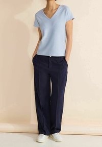 Ljust blå randig kortärmad V-hals topp matchad med löst sittande marinblå byxor och vita sneakers, mot en neutral bakgrund.