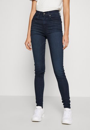 Jean skinny taille haute bleu foncé en denim avec une texture lisse, présentant une fermeture à bouton à l'avant, cinq poches et un design ajusté.