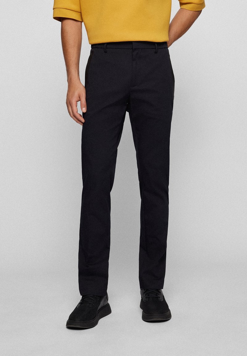 Homme portant un pantalon noir droit, des chaussures noires et une chemise jaune moutarde à manches longues, debout devant un fond uni.