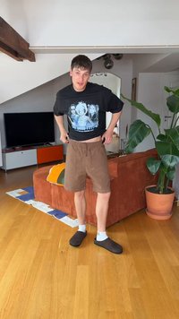 Jeune homme ajuste un short marron à l'intérieur, portant un t-shirt noir graphique, des chaussettes blanches et des chaussons foncés, debout sur un sol en bois près d'un canapé et d'une plante.