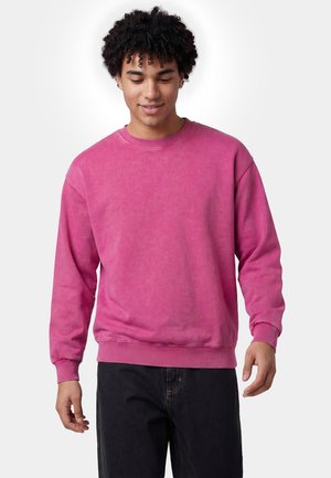 Junger Mann mit lockigem Haar, der ein hellpinkes langärmliges Sweatshirt und schwarze Hose trägt und mit einem leichten Lächeln nach unten schaut.