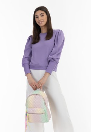 Lila Sweatshirt mit Puffärmeln; pastellfarbener, gradient-quilteter Rucksack mit Reißverschluss und bunten Quasten; Model trägt weiße Hosen.
